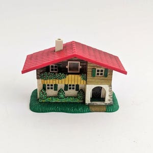 Puede incluir: Una casa decorativa en miniatura con tejado rojo, contraventanas verdes y chimenea. La casa está sobre una base verde, y la fachada presenta una pequeña puerta arqueada y múltiples ventanas. El diseño general evoca una estética pintoresca y rústica.