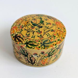 May include: A round, yellow lidded box featuring a hand-painted design of various birds, green palm-like foliage, and small red floral accents. The cylindrical container has a smooth finish and a repeating botanical pattern across the lid and sides.