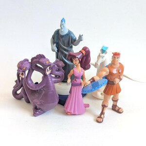 May include: A collection of Hercules movie figurines. The set includes Hercules, Megara, Hades, a three-headed Hydra, and a white Pegasus. The figurines are brightly colored and made of plastic. The characters are posed in action.