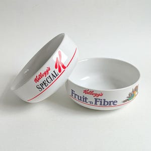 Puede incluir: Dos cuencos de cerámica blanca para cereales. Uno tiene el texto "Kellogg's SPECIAL" en rojo y la letra "R" en rojo. El otro tiene el texto "Kellogg's Fruit 'n Fibre" en azul y rojo, con una ilustración colorida.