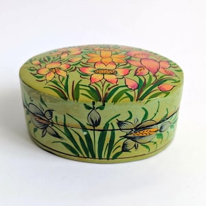 May include: A round, light green lidded box featuring hand-painted floral motifs in shades of orange, yellow, and dark green. The lid displays a cluster of blooming flowers, while the sides are decorated with smaller floral buds and green leaves.
