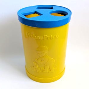 Peut inclure: Un trieur de formes cylindrique en plastique jaune avec un couvercle bleu comportant une ouverture carrée et une circulaire. Le côté du récipient jaune affiche le logo Fisher-Price en relief et un motif d'un enfant jouant avec des blocs.