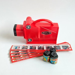 May include: A vintage red projector with the word "PROJECTOR" printed on the side. The image includes several color slides and film canisters. The slides and canisters have colorful illustrations. The text "MADE IN HONG KONG" is visible.