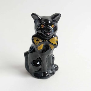May include: A glossy black ceramic cat figurine with yellow eyes and a yellow bow tie. The cat is sitting upright with its ears perked up. A decorative item for a shelf or desk.