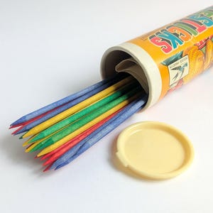 Puede incluir: Un juego de palillos chinos vintage. El recipiente cilíndrico es naranja con la palabra "STICKS" impresa. Los palillos son rojos, azules, amarillos y verdes. La tapa está a un lado.