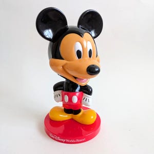 May include: A Mickey Mouse figurine with a black head and ears, an orange face, and a black nose. The figure wears a black body, red shorts with white buttons, yellow shoes, and white gloves. The figurine stands on a red base with "Walt Disney World Resort" printed on it.