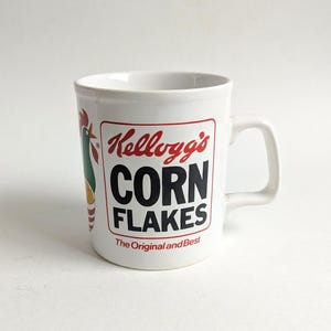 Op de afbeelding: Witte keramische mok met het Kellogg's Corn Flakes logo in rood en zwart. De mok heeft een oor en een hanenafbeelding aan de zijkant. De tekst "The Original and Best" staat onder het logo.