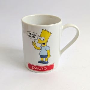 Könnte beinhalten: Weißer Keramikbecher mit Bart Simpson aus der Zeichentrickserie Die Simpsons. Bart zeigt das Peace-Zeichen, und der Becher trägt den Text "PEACE, MAN." und den Namen "DAVID" auf einem roten Banner. Der Becher hat einen gebogenen Henkel.