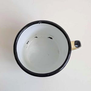 May include: White enamel mug with a black rim and handle. The mug has a black handle and a white interior. The mug is empty and sits on a white surface. The mug is a vintage style.