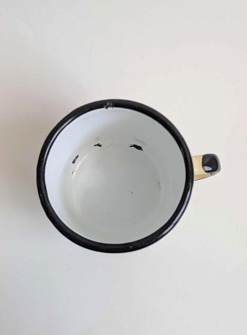 May include: White enamel mug with a black rim and handle. The mug has a black handle and a white interior. The mug is empty and sits on a white surface. The mug is a vintage style.