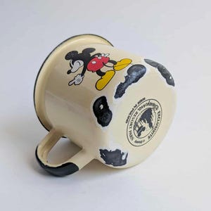 May include: Cream-colored enamel mug featuring Mickey Mouse in a classic pose. The mug has black accents on the handle and rim, with distressed black spots. The bottom of the mug has a Disney Resort Paris logo.