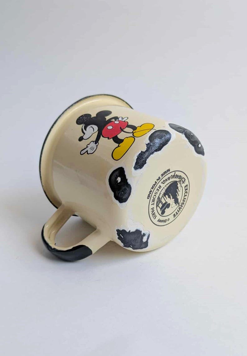 May include: Cream-colored enamel mug featuring Mickey Mouse in a classic pose. The mug has black accents on the handle and rim, with distressed black spots. The bottom of the mug has a Disney Resort Paris logo.