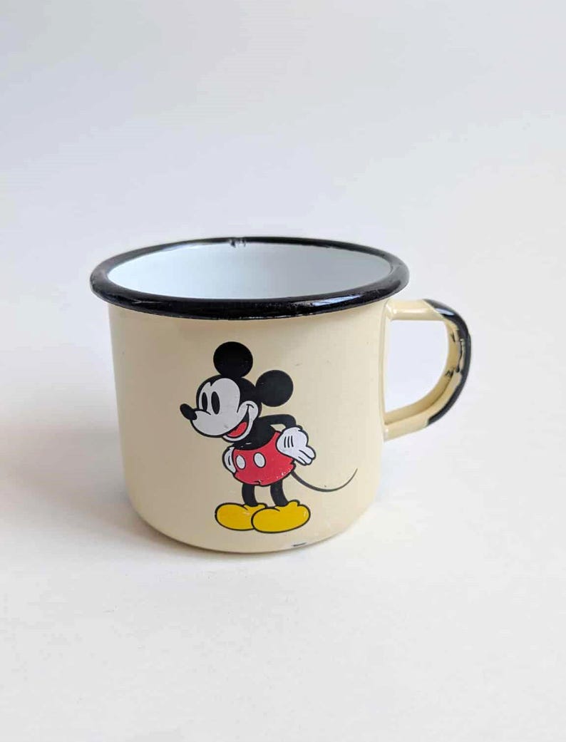 May include: Cream-colored enamel mug featuring a classic Mickey Mouse design. The mug has a black rim and handle, with Mickey in his iconic red shorts, yellow shoes, and white gloves. A vintage-style collectible.