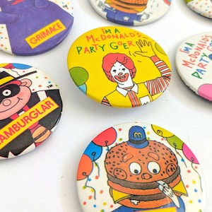 Op de afbeelding: Een verzameling vintage McDonald's feestbuttons met verschillende personages en zinnen. De buttons tonen Grimace, de Hamburglar, Ronald McDonald en een Big Mac-personage. Elke button is rond met kleurrijke illustraties en tekst.