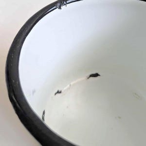 May include: Close-up of a white enamel bowl with a black rim. The bowl has some wear and tear, with small chips and imperfections visible on the surface. The interior is smooth and white.