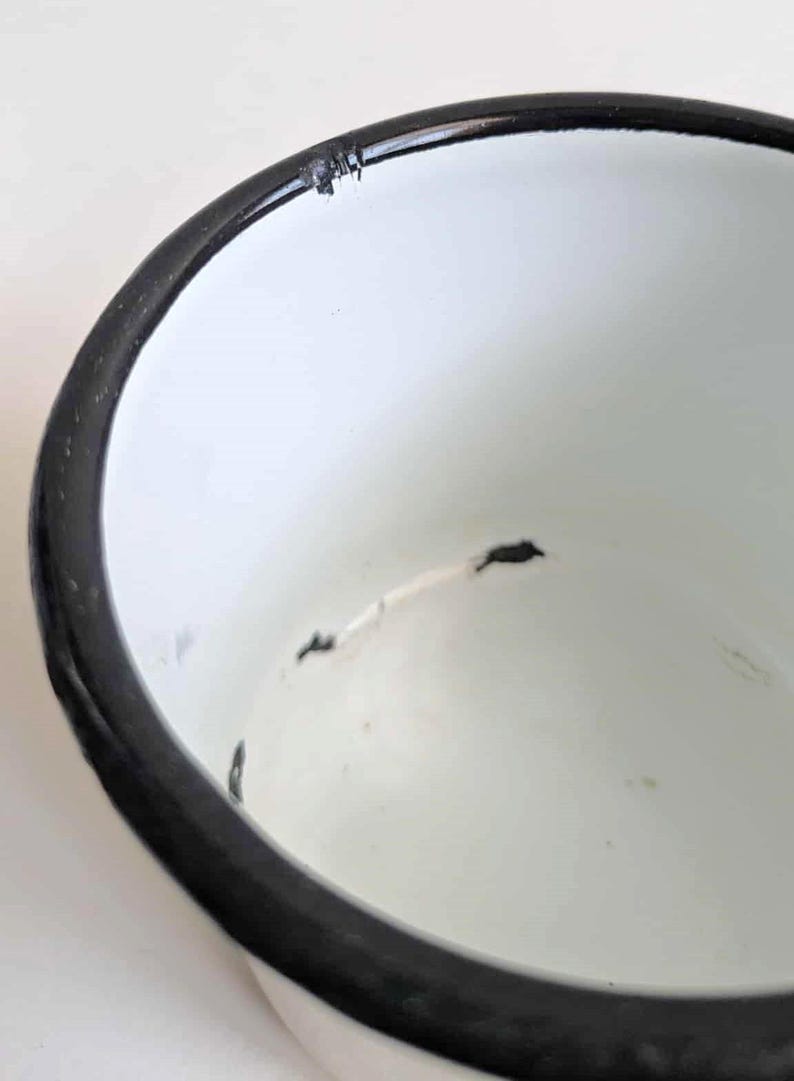 May include: Close-up of a white enamel bowl with a black rim. The bowl has some wear and tear, with small chips and imperfections visible on the surface. The interior is smooth and white.