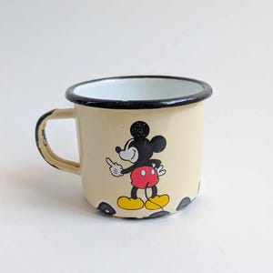 May include: Cream-colored enamel mug featuring a classic Mickey Mouse graphic. The mug has a black rim and handle, with black polka dots at the base. Mickey is depicted in his iconic red shorts and yellow shoes.