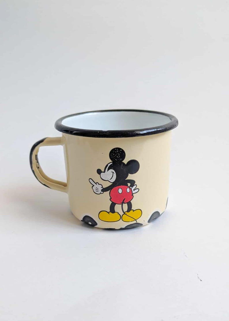 May include: Cream-colored enamel mug featuring a classic Mickey Mouse graphic. The mug has a black rim and handle, with black polka dots at the base. Mickey is depicted in his iconic red shorts and yellow shoes.