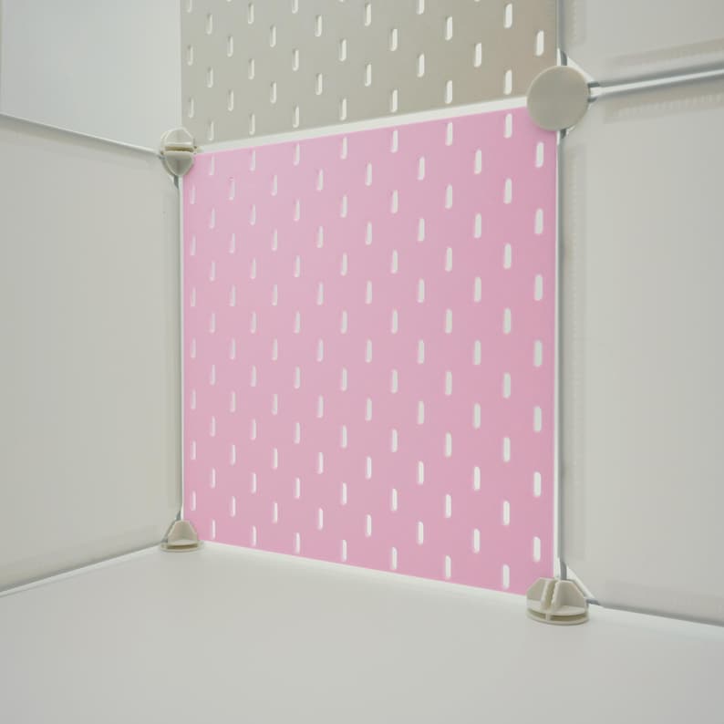 May include: A square pink pegboard panel featuring a repeating pattern of vertical slots. The panel is secured within a modular white frame system using light gray plastic corner connectors and circular fasteners at the edges of the structure.