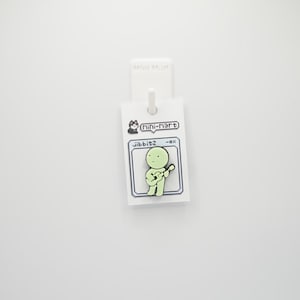 Puede incluir: Un pin verde claro con una figura de dibujos animados tocando la guitarra. El pin est&aacute; sujeto a una etiqueta blanca con el texto "Mini Mart" y "jibbitz -ox". El fondo es blanco liso.