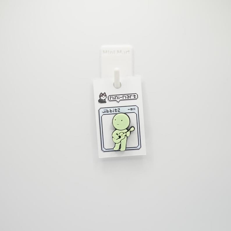 Puede incluir: Un pin verde claro con una figura de dibujos animados tocando la guitarra. El pin est&aacute; sujeto a una etiqueta blanca con el texto "Mini Mart" y "jibbitz -ox". El fondo es blanco liso.