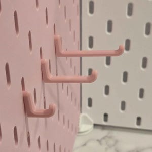 May include: Close-up of a pink pegboard with three pink hooks attached. The pegboard has evenly spaced oval holes. A white pegboard is visible in the background.
