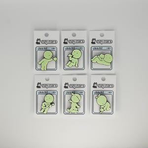 Puede incluir: Seis dijes Jibbitz verdes, cada uno con una figura de dibujos animados en diferentes poses. Los dijes est&aacute;n adheridos a tarjetas blancas con el texto "Mini-Mart" y "Jibbitz -ox". El fondo es blanco.