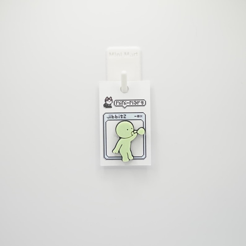 Puede incluir: Un charm jibbitz verde claro con la forma de una persona soplando una burbuja. El charm est&aacute; sujeto a una etiqueta blanca con el texto "(Mini-Mart)" y "jibbitz -0x". El fondo es blanco liso.