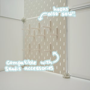 May include: A beige pegboard panel featuring vertical slots and multiple attached hooks. The panel includes white text overlays stating hooks also sold and compatible with Skadis accessories. The structure is supported by white corner connectors.
