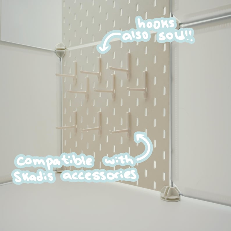 May include: A beige pegboard panel featuring vertical slots and multiple attached hooks. The panel includes white text overlays stating hooks also sold and compatible with Skadis accessories. The structure is supported by white corner connectors.