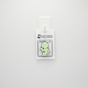 Puede incluir: Una etiqueta blanca con una imagen de dibujos animados de una figura verde claro, el texto "jibbitz -ox" y "mini-mart". La etiqueta est&aacute; sujeta a un gancho blanco.