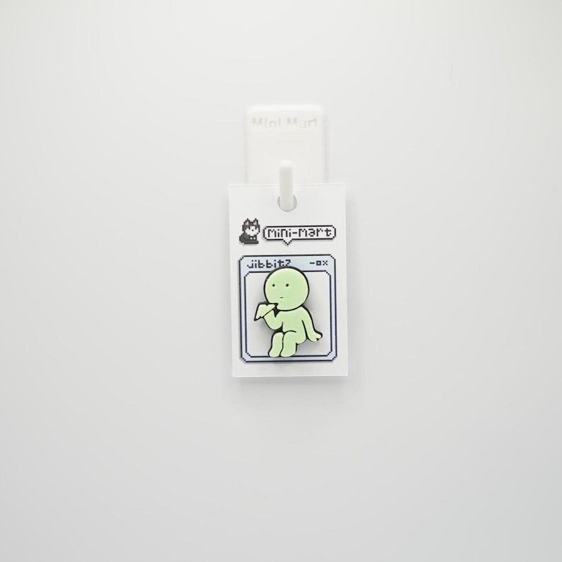 Puede incluir: Una etiqueta blanca con una imagen de dibujos animados de una figura verde claro, el texto "jibbitz -ox" y "mini-mart". La etiqueta est&aacute; sujeta a un gancho blanco.