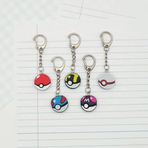 May include: Five circular keychains featuring various spherical designs with metallic silver chains and lobster claw clasps. The designs include a red and white ball, a yellow and black ball, a white and red ball, a blue and red ball, and a purple ball with an M.