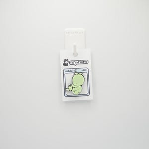 Puede incluir: Un charm jibbitz verde claro con un dise&ntilde;o de personaje de dibujos animados pixelado. El charm est&aacute; unido a una etiqueta blanca con el texto "Mini-Mart" y "jibbitz".