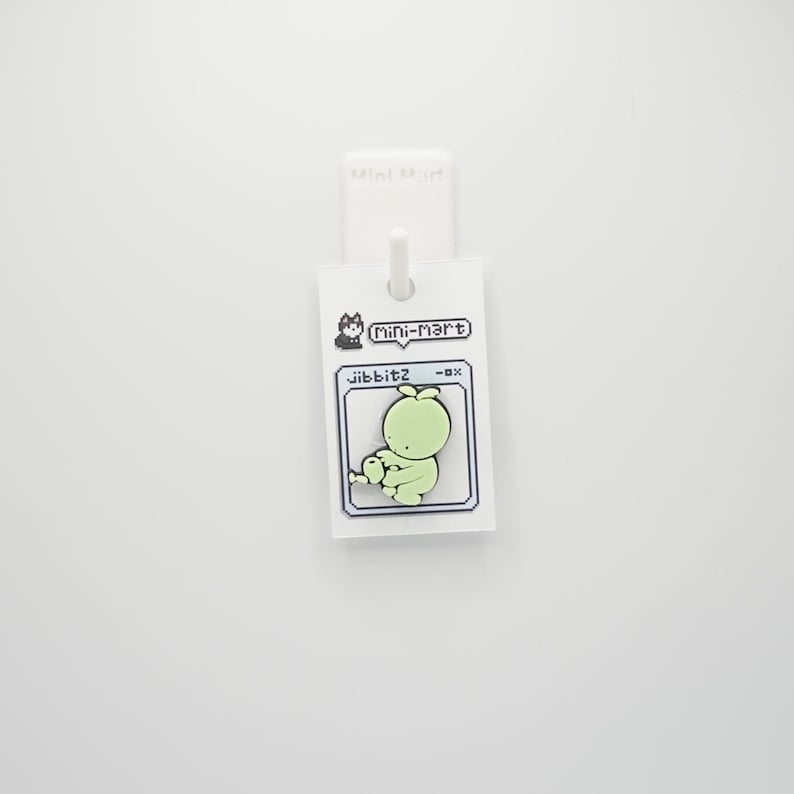 Puede incluir: Un charm jibbitz verde claro con un dise&ntilde;o de personaje de dibujos animados pixelado. El charm est&aacute; unido a una etiqueta blanca con el texto "Mini-Mart" y "jibbitz".