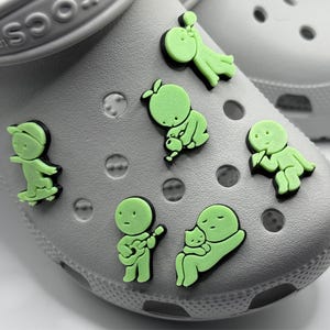 Puede incluir: Primer plano de zapatos Crocs grises adornados con varios dijes de zapatos verde ne&oacute;n. Los dijes representan varios personajes de dibujos animados, incluyendo un patinador, un m&uacute;sico y una figura sosteniendo un beb&eacute;.