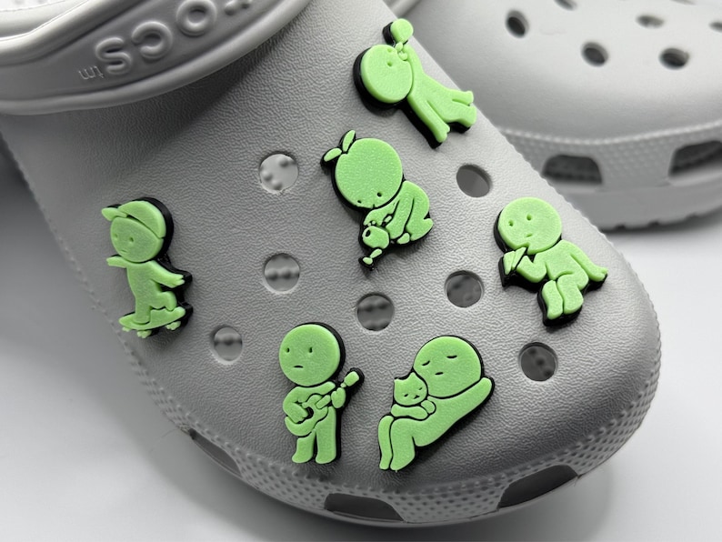 Puede incluir: Primer plano de zapatos Crocs grises adornados con varios dijes de zapatos verde ne&oacute;n. Los dijes representan varios personajes de dibujos animados, incluyendo un patinador, un m&uacute;sico y una figura sosteniendo un beb&eacute;.
