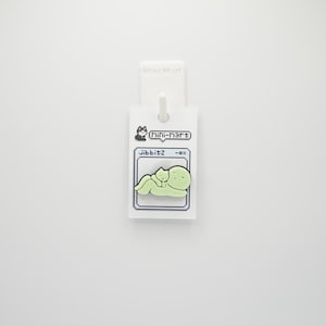 Puede incluir: Un charm Jibbitz verde claro con un gato de dibujos animados y una figura redondeada m&aacute;s grande. El charm est&aacute; unido a una etiqueta blanca con el texto "Mini-mart" y "Jibbitz -ox".