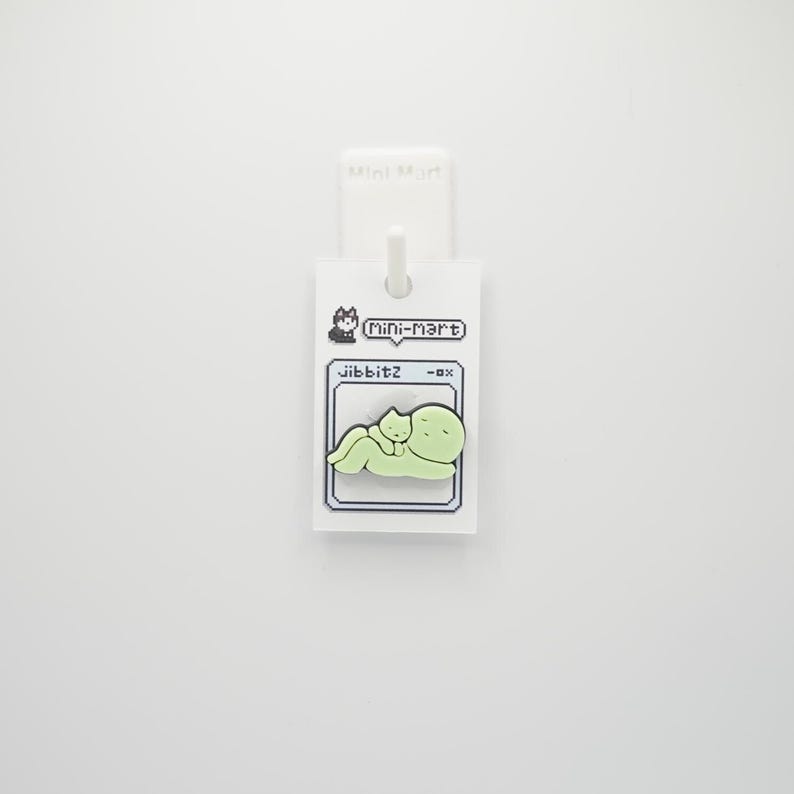 Puede incluir: Un charm Jibbitz verde claro con un gato de dibujos animados y una figura redondeada m&aacute;s grande. El charm est&aacute; unido a una etiqueta blanca con el texto "Mini-mart" y "Jibbitz -ox".