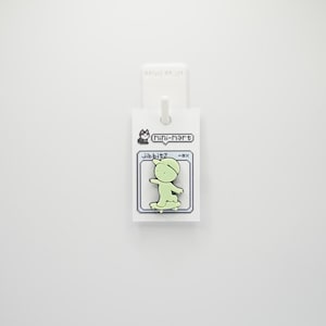 Puede incluir: Un charm Jibbitz verde claro con un personaje de dibujos animados montando en monopat&iacute;n. El charm est&aacute; unido a una etiqueta blanca con el texto "Mini-Mart" y "Jibbitz". El dise&ntilde;o tiene una est&eacute;tica pixelada.