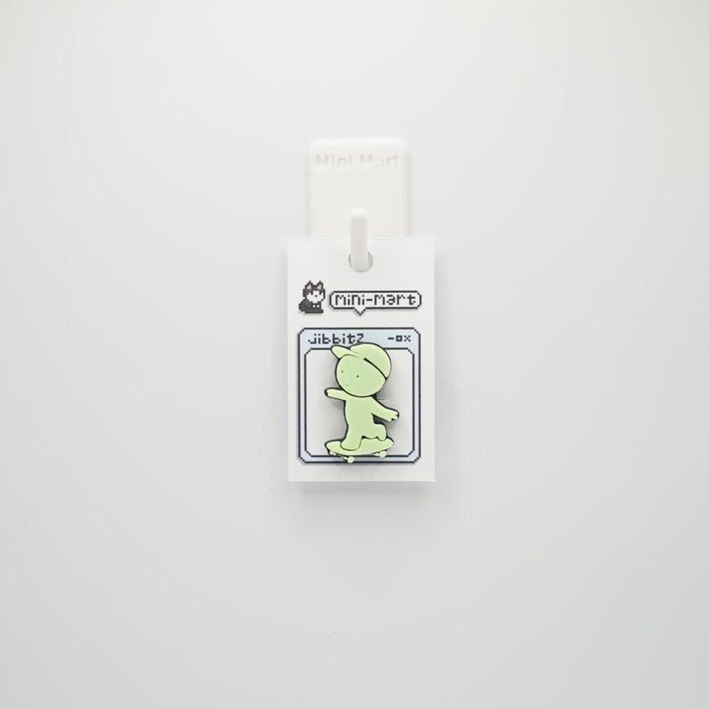 Puede incluir: Un charm Jibbitz verde claro con un personaje de dibujos animados montando en monopat&iacute;n. El charm est&aacute; unido a una etiqueta blanca con el texto "Mini-Mart" y "Jibbitz". El dise&ntilde;o tiene una est&eacute;tica pixelada.