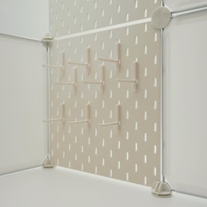 May include: A beige pegboard panel featuring a grid of vertical slots and multiple attached L-shaped hooks. The panel is secured within a white modular frame structure with circular and triangular corner connectors, creating a storage display.