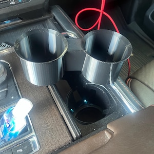 May include: Black 3D-printed car cup holder insert with two angled cup holders. The insert sits in a car's center console, next to the existing cup holder. A red charging cable is visible.