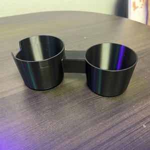 May include: A black dual cup holder accessory featuring two cylindrical compartments connected by a central bridge. The left compartment includes a small U-shaped cutout along the top rim, while both containers have smooth, tapered walls.