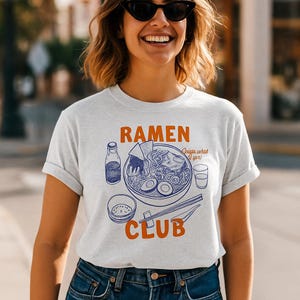 Puede incluir: Camiseta gris claro con un diseño de un cuenco de ramen, una botella y palillos. La palabra "RAMEN" está en naranja encima del cuenco, y "CLUB" debajo. La frase "...what it'yor!" está a la derecha.