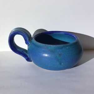 May include: A small, rounded ceramic mug featuring a vibrant blue glaze with subtle variations in tone. The mug has a thick, curved handle attached to one side and a smooth, glossy finish that reflects light, showing a deep blue interior rim.