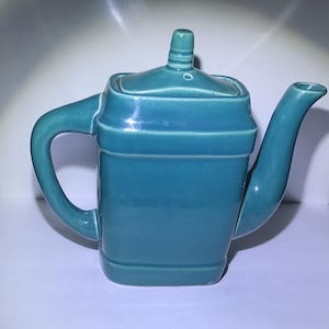 May include: A teal ceramic teapot with a square-shaped body, a curved handle on the left side, and a long, slender spout on the right. The teapot features a matching square lid with a small, central knob handle on top, all finished in a smooth, glossy glaze.