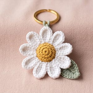 Può includere: Un portachiavi a margherita all'uncinetto con un disco centrale circolare giallo senape circondato da petali bianchi. Una piccola foglia all'uncinetto color verde salvia è attaccata al lato del fiore. Il fiore è collegato a un anello in metallo dorato.