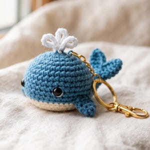 以下が含まれることがあります： 青いかぎ針編みのクジラのキーホルダー。頭頂部には白い糸で作られた潮吹きがあり、黒いプラスチック製のビーズの目とクリーム色の腹部が特徴です。クジラは金色の金属製キーリングとナスカンに取り付けられており、細部まで丁寧に編み込まれています。