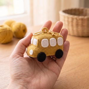 Könnte beinhalten: Ein gelber, gehäkelter Schulbus-Schlüsselanhänger mit weißen, runden Fenstern, schwarzen Rädern und einem goldenen Karabinerverschluss. Der Bus ist in einer strukturierten Häkeloptik gefertigt und verfügt über einen kleinen Frontscheinwerfer.